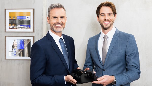 Двоє ділових людей стоять у приміщенні та посміхаються в камеру.