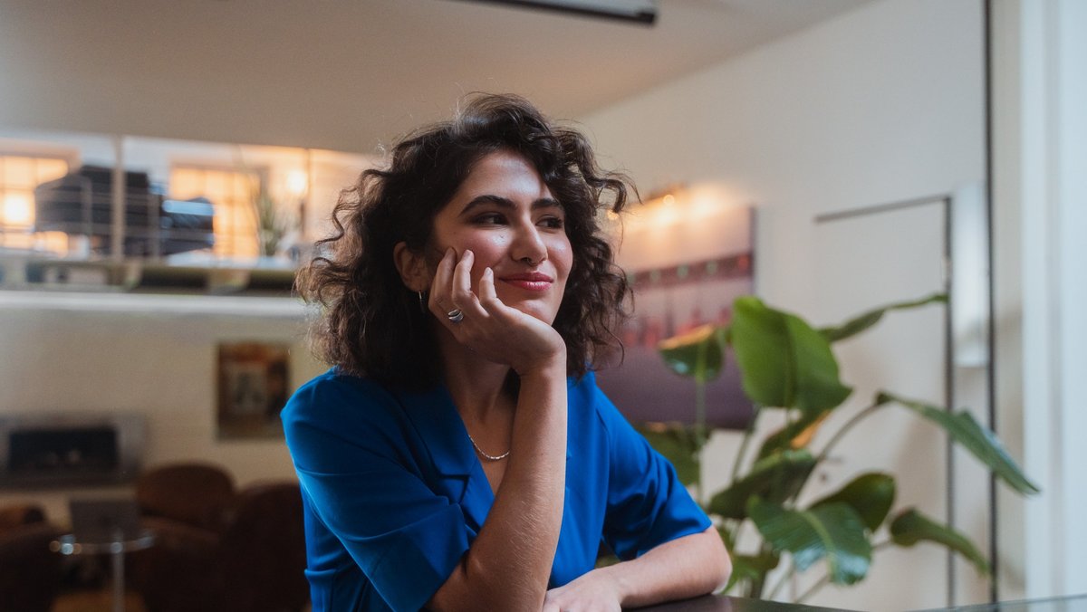 Жінка сидить у приміщенні та з усмішкою задумливо дивиться у вікно.