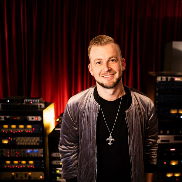 Усміхнений чоловік стоїть у студії звукозапису з аудіообладнанням на тлі.