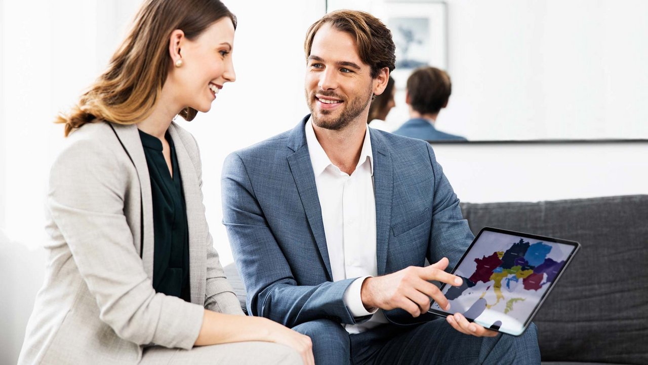 Чоловік і жінка сидять разом, посміхаються та дивляться на планшет у професійному середовищі.