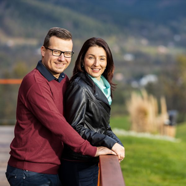 Усміхнені чоловік і жінка стоять на відкритому просторі, спираючись на перила, на тлі пейзажу.