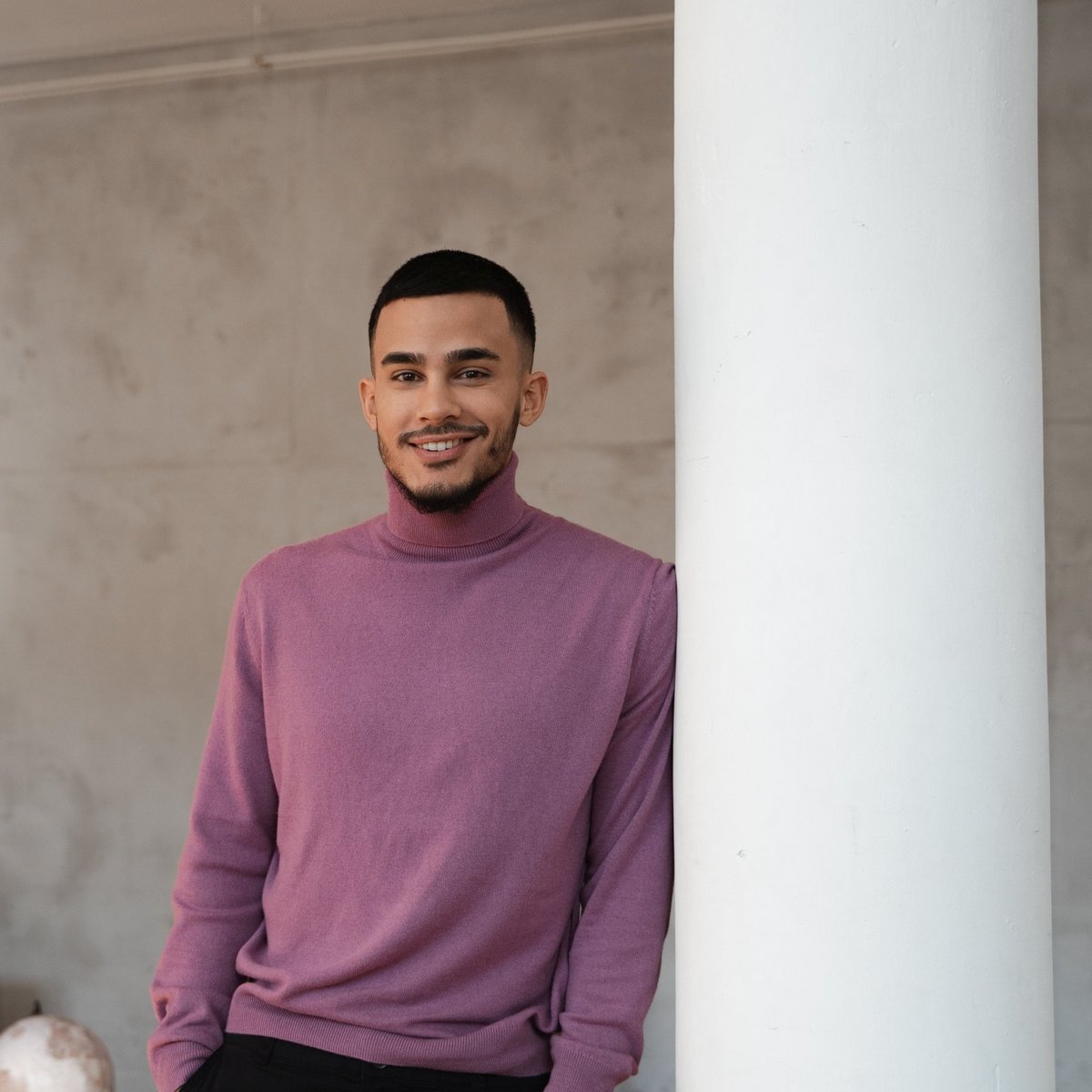 Чоловік стоїть у приміщенні та усміхається біля колони.